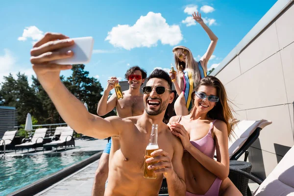 Arabian man with beer taking selfie with friends in swimwear near swimming pool — Stock Photo