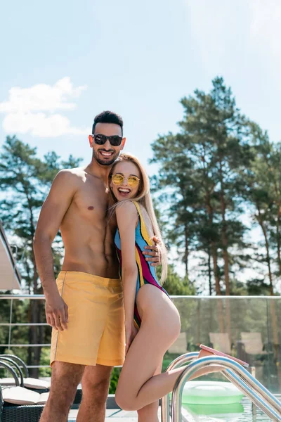 Arabian man in swimming trunks hugging friend in swimsuit and sunglasses near swimming pool — Stock Photo