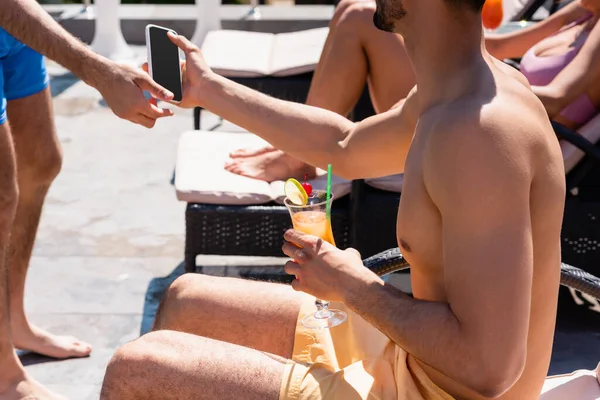Cropped view of man giving smartphone to friend in swimming trunks with cocktail — Stock Photo