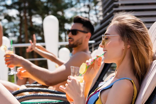 Smiling woman in swimsuit drinking cocktail near blurred friend at resort — Stock Photo