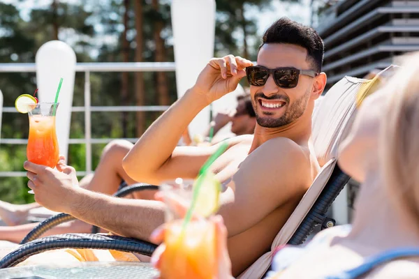 Fröhlicher arabischer Mann mit Sonnenbrille hält Cocktail neben verschwommenem Freund auf Liegestuhl — Stockfoto