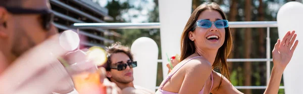 Усміхнена жінка махає рукою біля розмитих друзів на курорті, банер — стокове фото