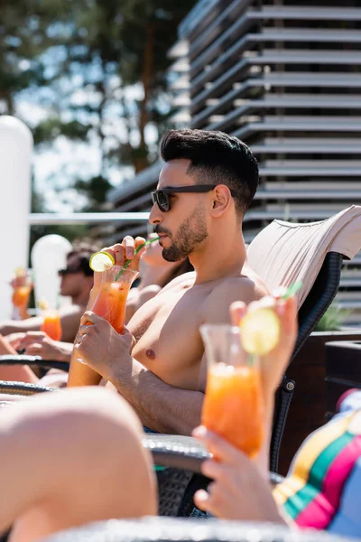 Arabian man in sunglasses drinking cocktail near blurred friend in swimsuit — Stock Photo