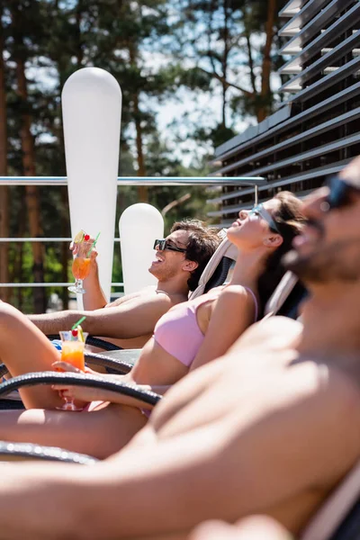 Vue latérale d'amis joyeux avec des cocktails allongés sur des chaises longues à l'extérieur — Photo de stock