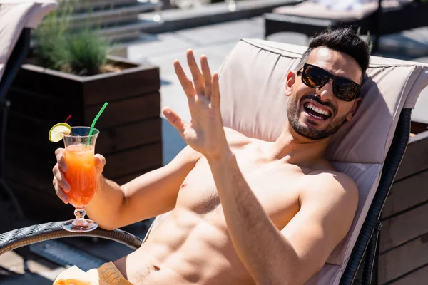 Happy muslim man in swimming trunks waving hand and holding cocktail on deck chair — Stock Photo