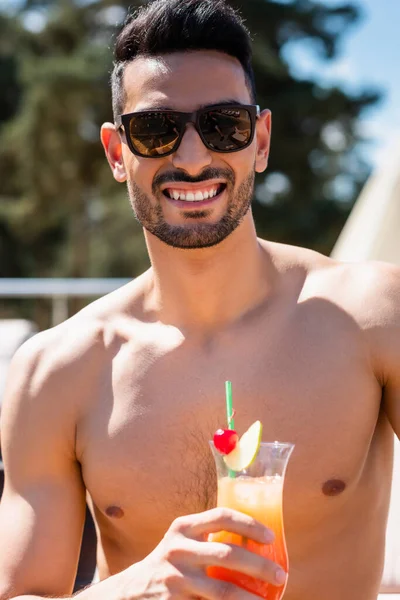 Sorrindo homem muçulmano sem camisa em óculos de sol segurando coquetel ao ar livre — Fotografia de Stock