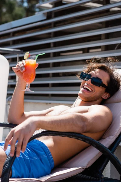 Homme positif dans les maillots de bain tenant un cocktail sur une chaise longue — Photo de stock