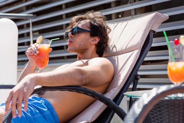 Mann mit Sonnenbrille hält Cocktail in der Hand, während er auf Liegestuhl im Resort relaxt — Stockfoto