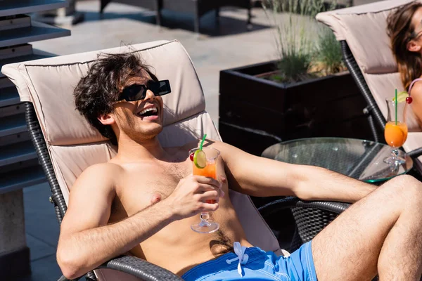 Positive man in swimming trunks and sunglasses holding cocktail at resort — Stock Photo