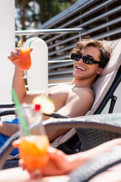 Smiling man in sunglasses and swimming trunks holding cocktail near blurred friend during weekend — Stock Photo