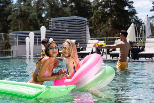 Donne eccitate ridendo della macchina fotografica mentre riposano in piscina vicino ad amici multietnici offuscati — Foto stock