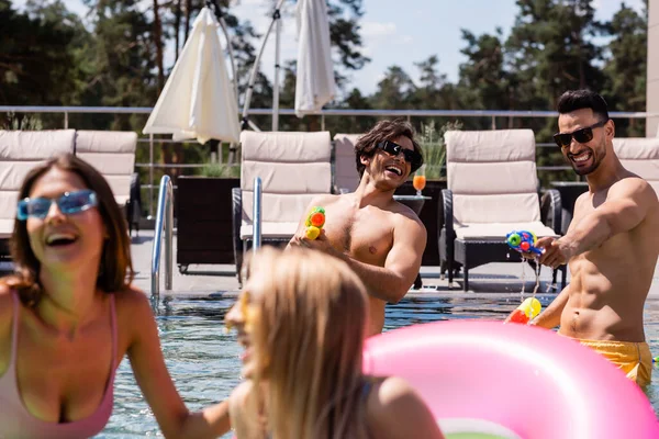 Веселі міжрасові чоловіки грають з водяними гарматами біля розмитих жінок у басейні — стокове фото