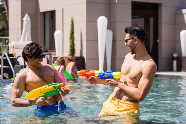 Animado interracial homens fazendo água pistolas batalha perto de desfocado mulheres no piscina — Fotografia de Stock