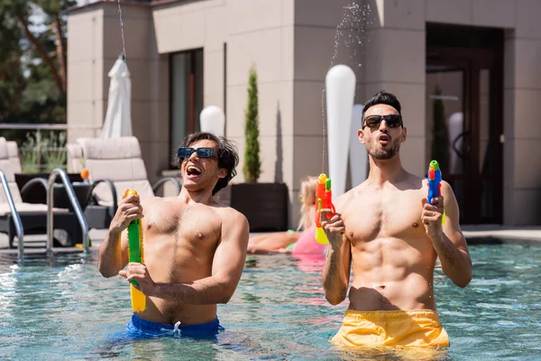 Homens multiculturais em calções de banho se divertindo enquanto brincavam com pistolas de água na piscina — Fotografia de Stock