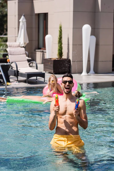 Aufgeregter muslimischer Mann spielt mit Wasserpistolen in der Nähe von Freunden im Pool — Stockfoto