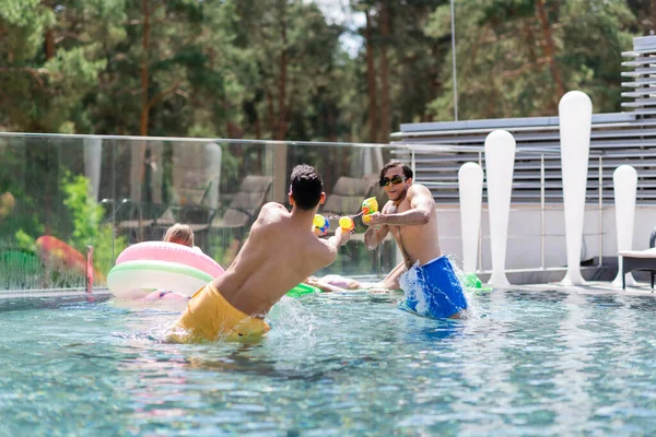 Young man having water pistols battle with arabian friend in swimming pool — Stock Photo