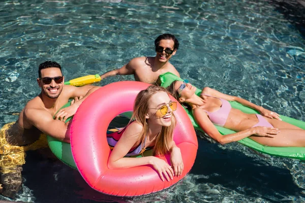 Cheerful woman laughing on swim ring in swimming pool near interracial friends — Stock Photo