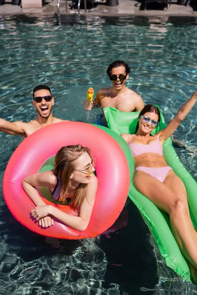 Donne felici su materasso gonfiabile e anello di nuoto a riposo in piscina vicino a uomini multietnici — Foto stock