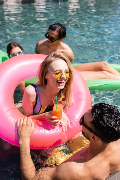 Mujer alegre con cóctel riendo en la piscina cerca de amigos multiculturales - foto de stock