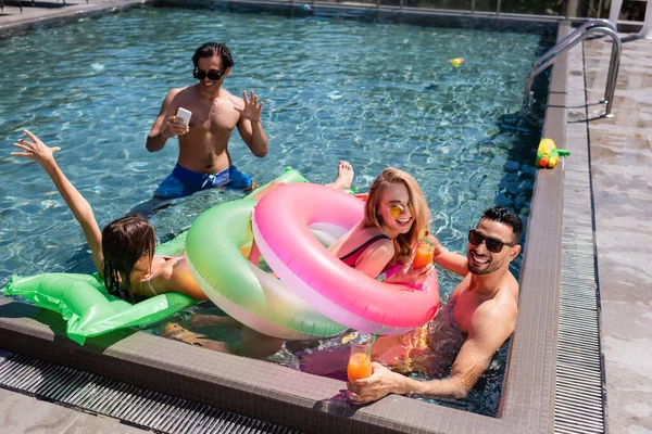 Homem feliz com a mão acenando smartphone perto de amigos inter-raciais descansando na piscina — Fotografia de Stock