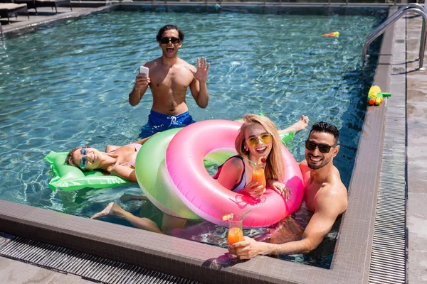 Hombre emocionado con teléfono inteligente saludando mano cerca de amigos multiétnicos en la piscina - foto de stock