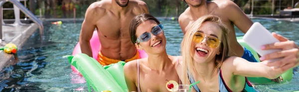 Возбужденные женщины делают селфи на смартфоне с многонациональными друзьями в бассейне, баннер — стоковое фото