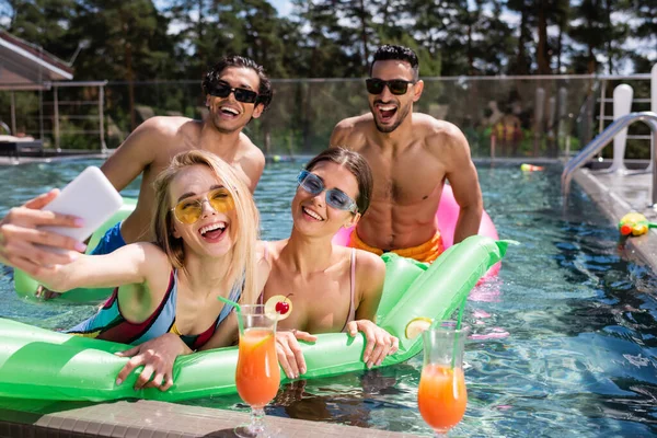 Mujer emocionada tomando selfie en la piscina cerca de cócteles y amigos interracial - foto de stock