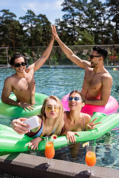 Feliz interracial los hombres dando alta cinco cerca de las mujeres tomando selfie en piscina - foto de stock