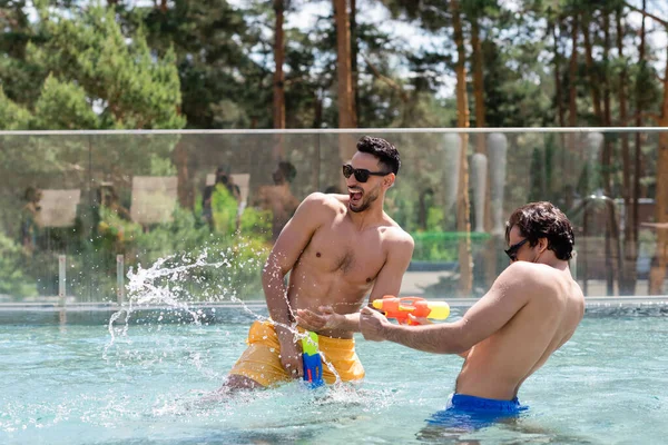 Alegre homem árabe em óculos de sol e calções de banho se divertindo com o amigo na piscina — Fotografia de Stock