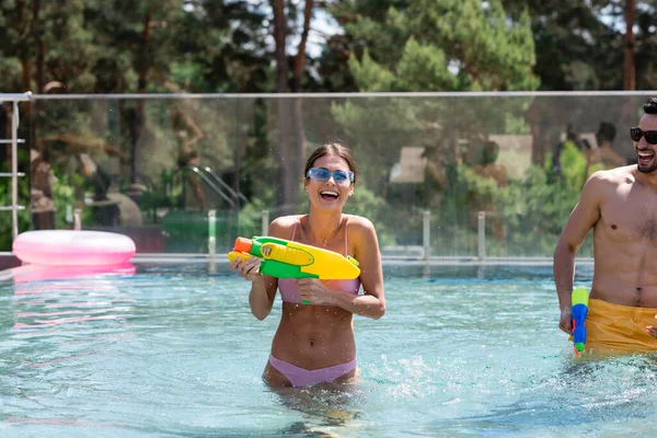 Молодая женщина с водяным пистолетом смеется в бассейне возле арабского мужчины — стоковое фото