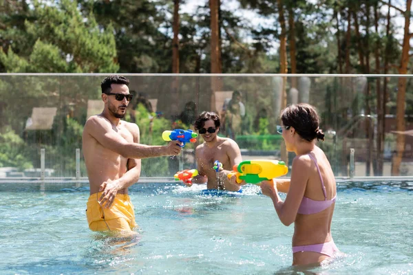 Amigos multiétnicos se divertindo durante batalha pistolas de água na piscina — Fotografia de Stock