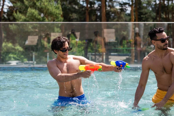 Joven en gafas de sol jugando con pistolas de agua cerca de amigo árabe en la piscina - foto de stock