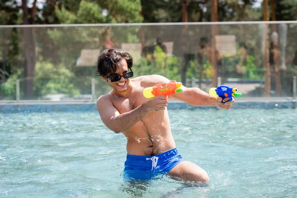 Aufgeregter Mann in Badehose und Sonnenbrille spielt mit Wasserpistolen im Pool — Stockfoto