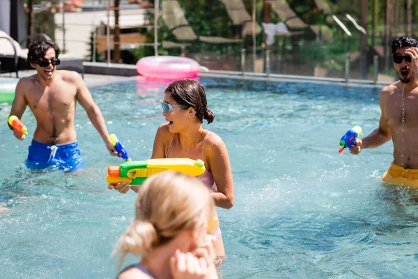 Cheerful interracial friends fighting on water guns in swimming pool — Stock Photo