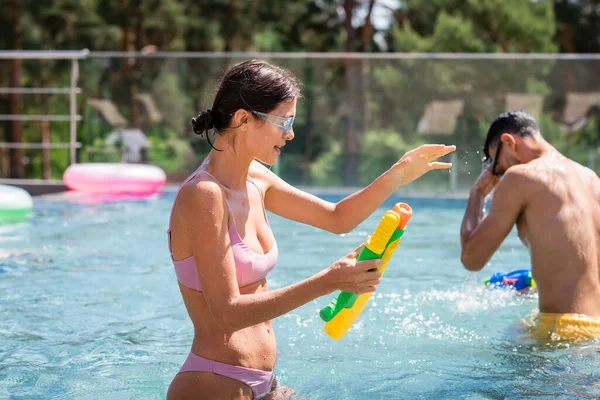 Young woman with water gun gesturing near arabian friend in pool — Stock Photo