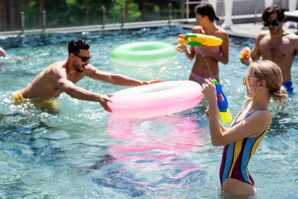 Multicultural friends having fun in swimming pool with swim rings and water guns — Stock Photo