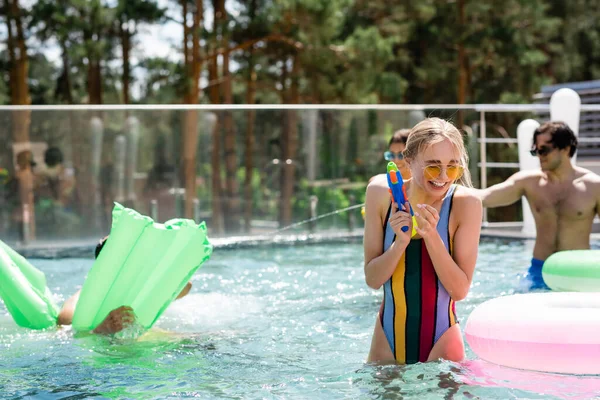 Cheerful woman in striped swimsuit holding water pistol near friends in pool — Stock Photo