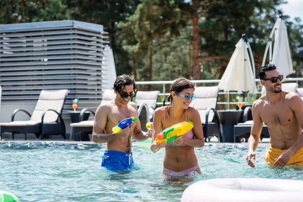 Happy young woman with interracial men playing with water guns in swimming pool — Stock Photo