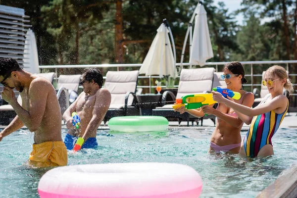 Aufgeregte Frauen kämpfen mit Wasserpistolen neben multiethnischen Freunden im Schwimmbad — Stockfoto