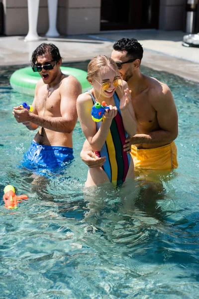 Amigos interracial alegre jugando con pistolas de agua en la piscina - foto de stock