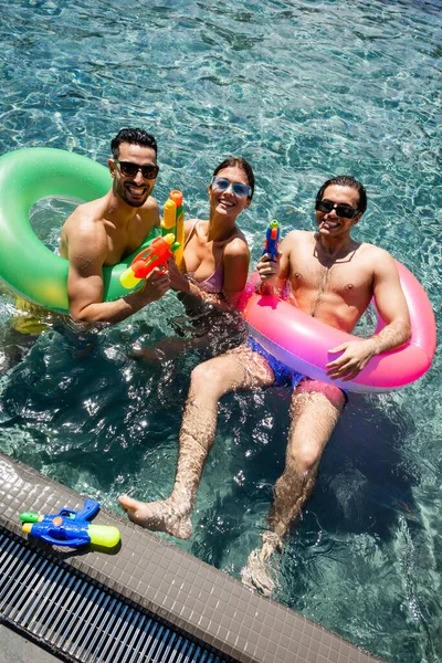 Vista de alto ângulo de felizes amigos inter-raciais com anéis de natação e armas de água na piscina — Fotografia de Stock