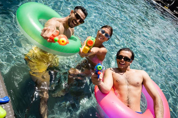 Захоплені багатоетнічні друзі розважаються з водяними пістолетами і плавають кільцями в басейні — стокове фото