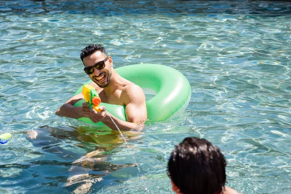 Animado árabe homem se divertindo com água pistola e anel de natação na piscina — Fotografia de Stock