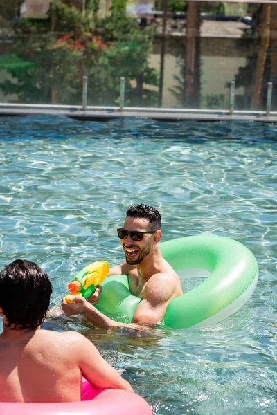 Alegre árabe homem em óculos de sol brincando com água arma perto amigo na piscina — Fotografia de Stock