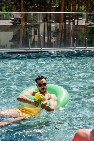 Animado muçulmano homem flutuando no anel de natação e jogando arma de água na piscina — Fotografia de Stock