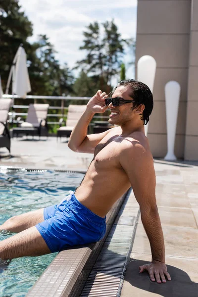 Hombre sonriente en bañador ajustando gafas de sol mientras está sentado cerca de la piscina - foto de stock