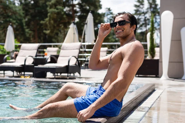 Homem alegre tocando óculos de sol e olhando para longe enquanto sentado à beira da piscina — Fotografia de Stock