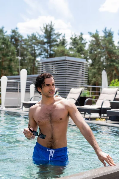 Hombre sin camisa sosteniendo gafas de sol y mirando en la piscina - foto de stock