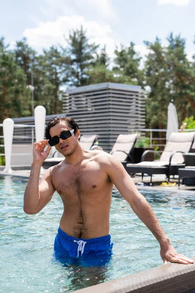 Hombre sin camisa en traje de baño de pie en la piscina y el ajuste de las gafas de sol - foto de stock