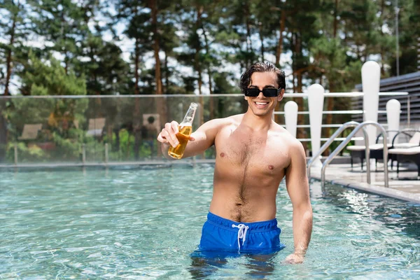 Мужчина без рубашки в солнцезащитных очках держит пиво во время отдыха на курорте — стоковое фото
